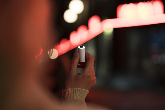 Hand hält eine moderne E-Zigarette vor verschwommenem Hintergrund mit roten Lichtern Nahaufnahme einer Hand, die eine E-Zigarette in der Dämmerung hält, vor einem unscharfen Hintergrund mit roten Leuchtreklamen