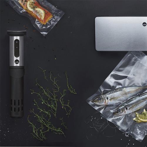 Sous-Vide Gargerät neben vakuumverpacktem Lachs und Fisch auf einem dunklen Untergrund mit frischen Kräutern