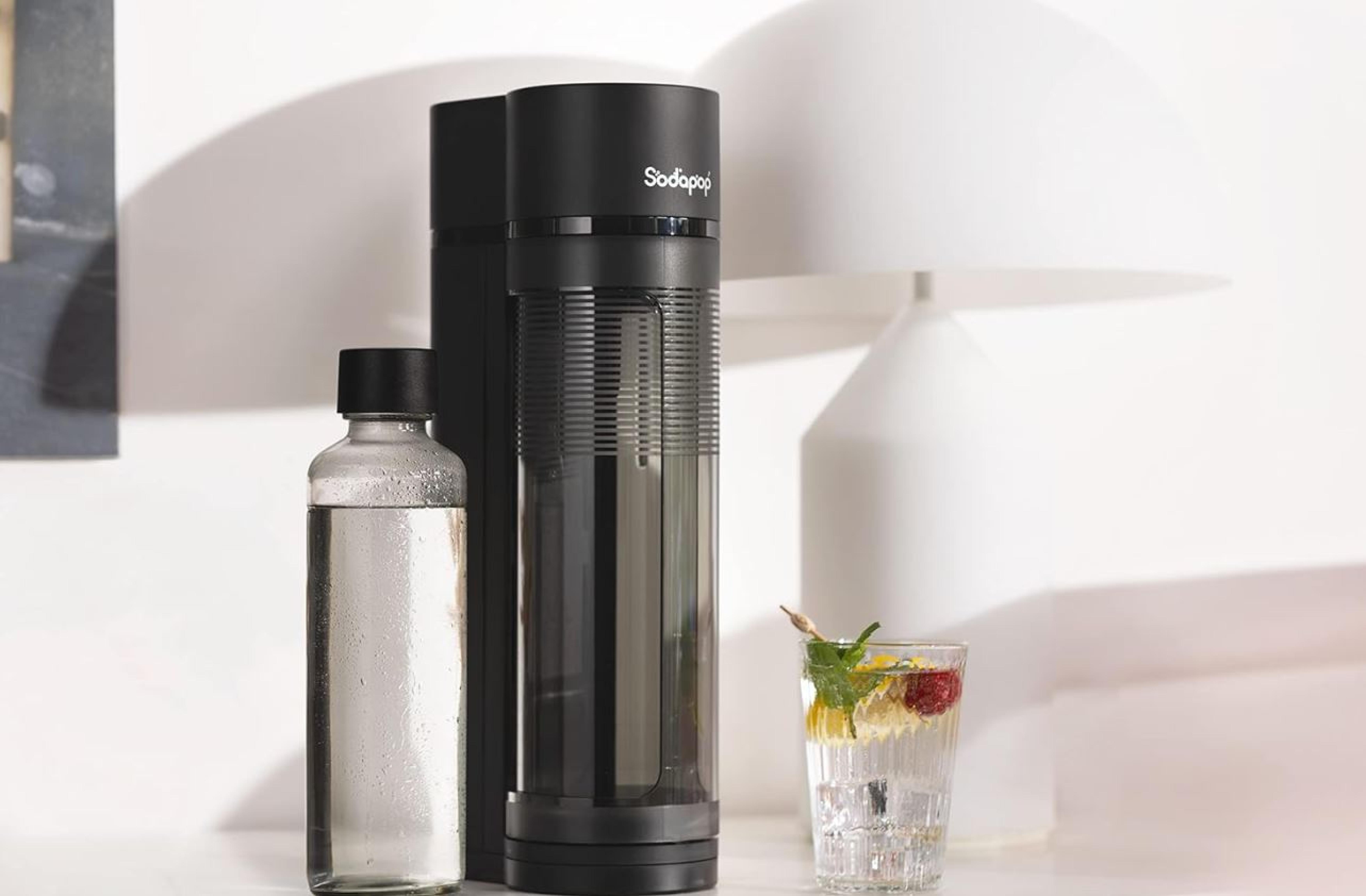 Ein schwarzer Wassersprudler mit Glasflasche neben einem Glas Wasser mit einer Zitronenscheibe und Beeren, auf einer hellen Oberfläche platziert.