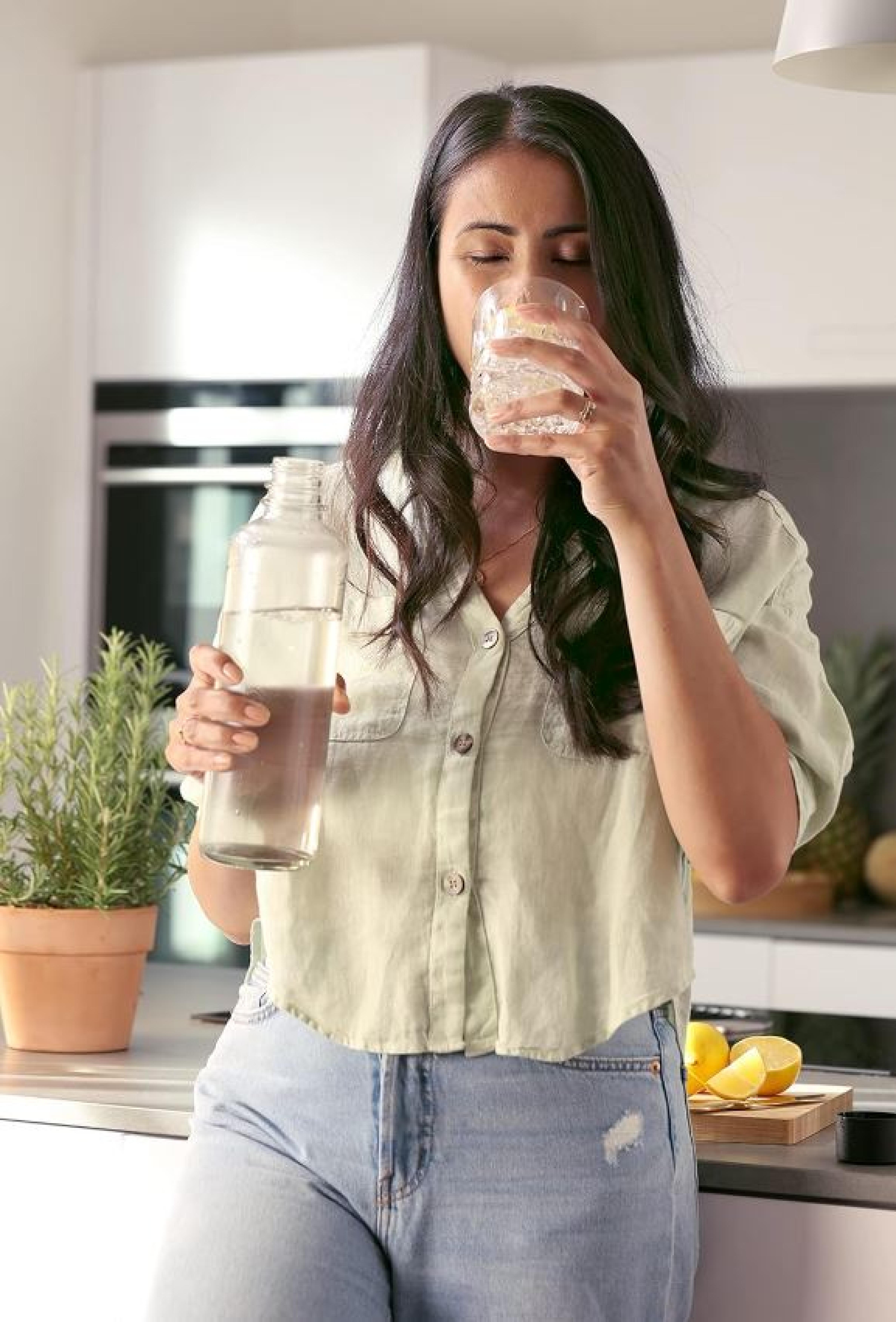 Junge Frau in heller Bluse und Jeans trinkt Wasser aus einem Glas und hält eine Wasserflasche in einer modernen Küche mit Pflanzen im Hintergrund.